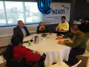 In this image, there are five adults conversing around a table with the NEADS banner behind one individual. 